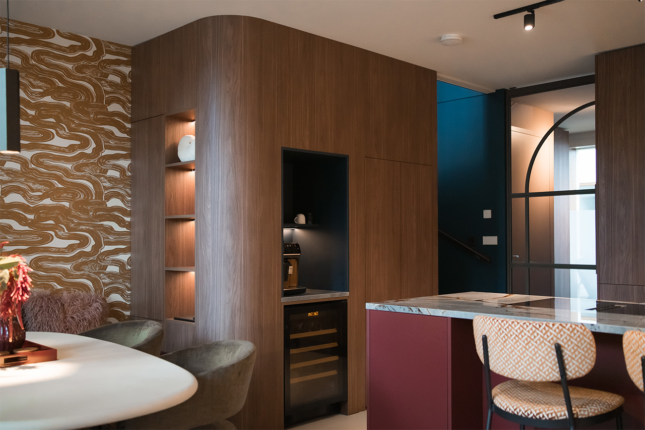 Moderne open keuken met houten kastenwand, ingebouwde wijnkoelkast en koffienis, kookeiland met marmeren blad en barstoelen, naast een ronde eettafel met designstoelen en sfeervolle verlichting. Interieuradvies in Alkmaar nodig? Neem contact op met Studio Kabel!